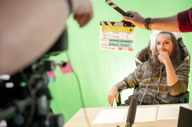 Regisseur sitzt konzentriert im Regiestuhl vor einem Greenscreen, im Vordergrund hält eine Person die Filmklappe zu „4 Profiles“